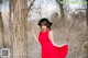 A woman in a red dress and black hat leaning against a tree.